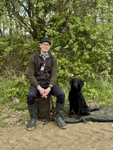 Sascha sitzt in Gummistiefeln am Wegesrand, neben ihm sitzt Femmy, ein schwarzer Labrador, auf einer Decke.